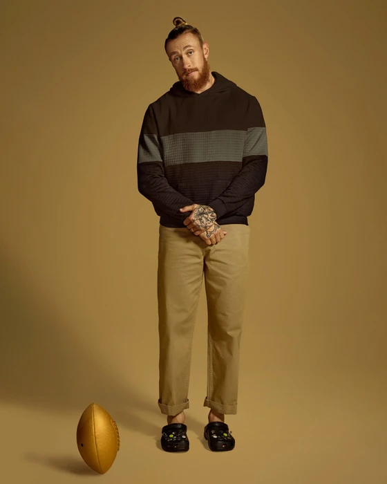 Man with reddish beard and topknot wearing dark hoodie and khaki trousers standing with a gold football