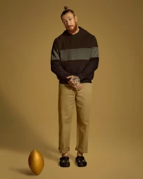 Man with reddish beard and topknot wearing dark hoodie and khaki trousers standing with a gold football