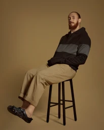 Man with red beard wearing black hoodie and tan trousers seated on a stool against a brown studio backdrop.