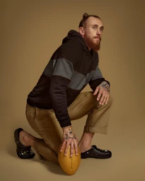 Man with tattoos and top knot kneeling in a black hoodie and khaki pants holding a gold football against a warm beige background.