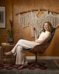 Woman with curly brown hair seated in a wooden armchair holding a red apple against a wood-paneled wall