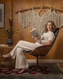 Woman in cream clothing seated in a mid-century modern wooden armchair holding a book in front of a wood-paneled wall with macrame wall hanging