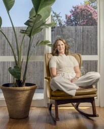 Woman with curly red hair seated cross-legged in a vintage armchair beside a large potted plant indoors.