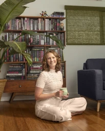 Woman sitting cross-legged on a wooden floor holding a mug in a living room with a bookshelf and blue sofa.