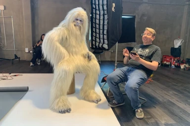 Behind-the-scenes photo by Kremer Johnson showing a person in a white furry Bigfoot costume seated next to a photographer in a studio.