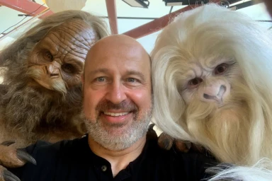 A smiling bald man taking a selfie flanked by two creature costume characters with detailed fur and masks