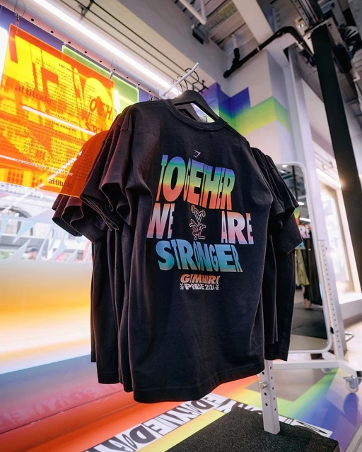 A black Gymshark t-shirt on a rack displays a colorful 'Together We Are Stronger' slogan in a retail setting with vibrant floor graphics.
