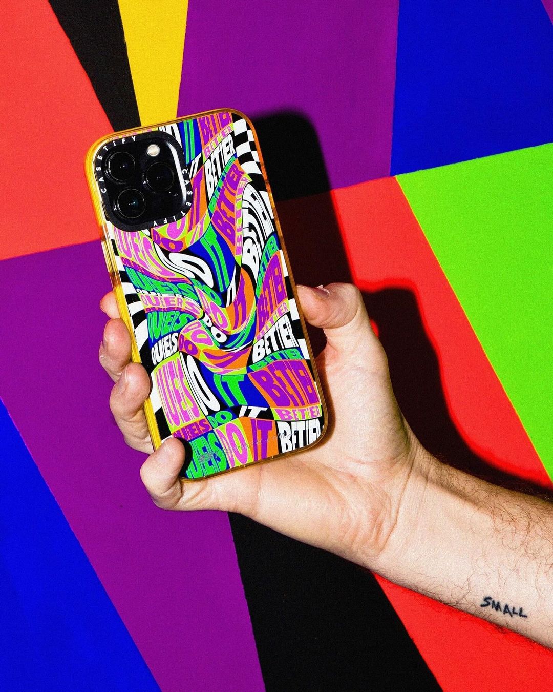 Hand holds a modern iPhone with a colorful patterned case against a vibrant, multi-colored geometric background of blue, red, purple, & green.