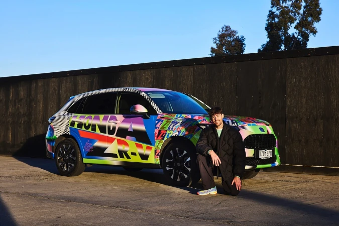 Man kneeling beside a vibrant, graffiti-style Honda CR-V on a concrete surface, set against a dark wooden fence and clear blue sky.