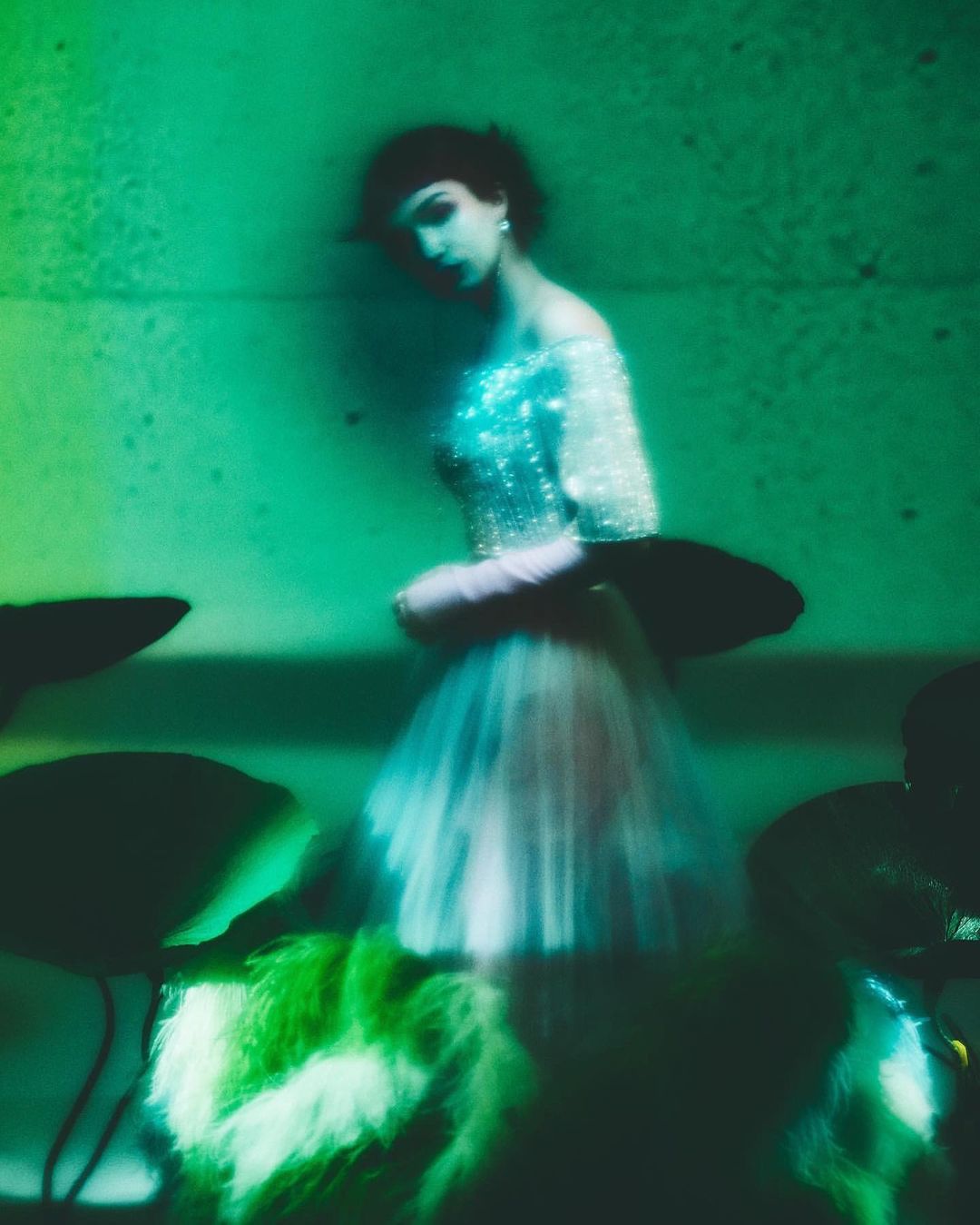 Elegant woman in shimmering gown poses indoors with intentional motion blur. She wears pale gloves and dark hair in a green-lit setting.