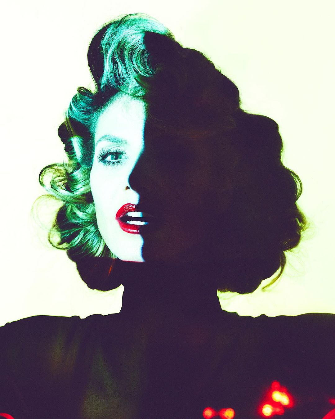 Portrait of a fair-skinned adult woman with red lipstick and stylized green hair, in shadows against a white background, medium shot.