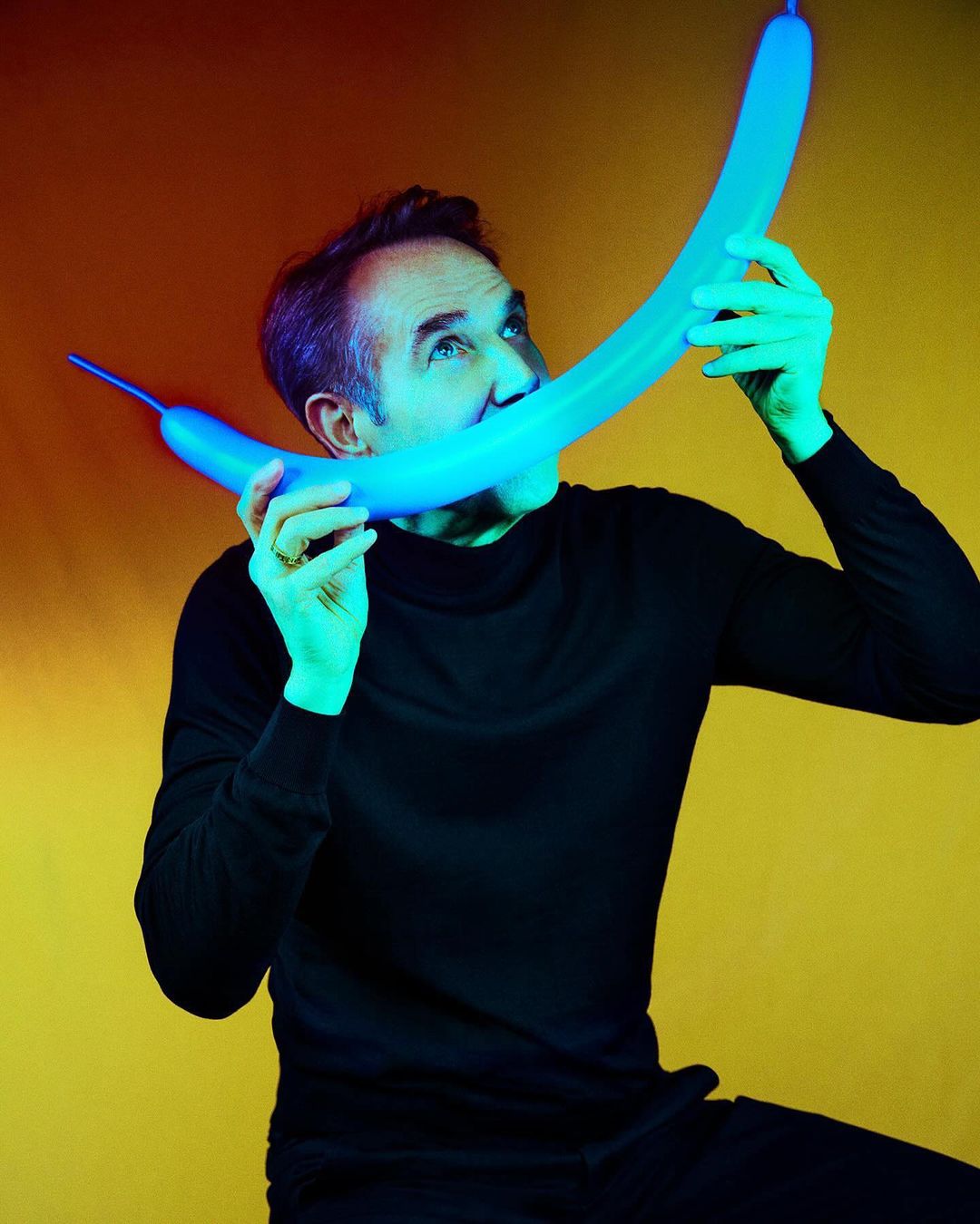 Adult man in black turtleneck poses with a blue balloon in his mouth against a yellow and orange studio backdrop with cyan lighting.