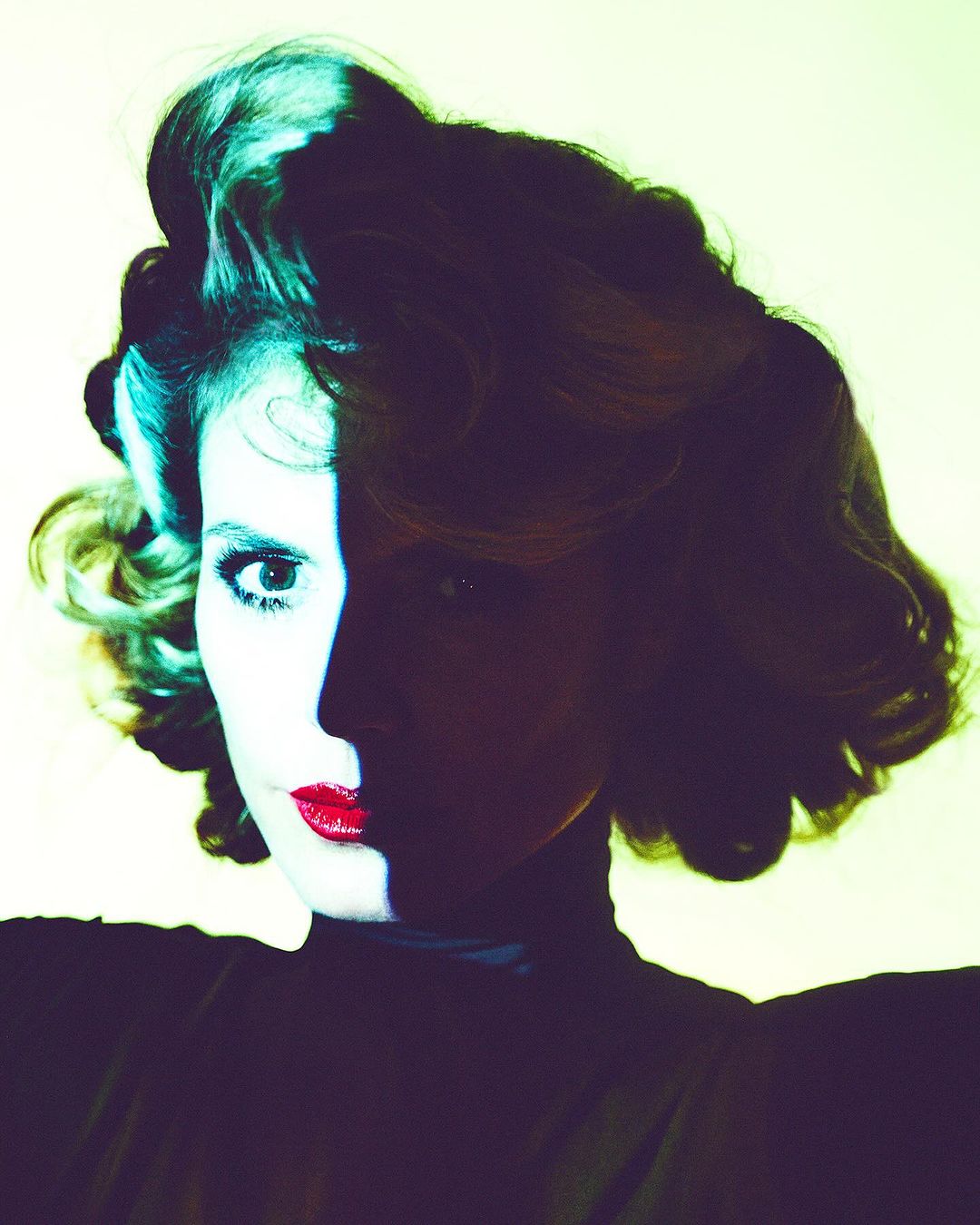 Dramatic portrait of a woman with retro hair, vivid red lips, and a face bisected by green-tinted light and deep shadow.
