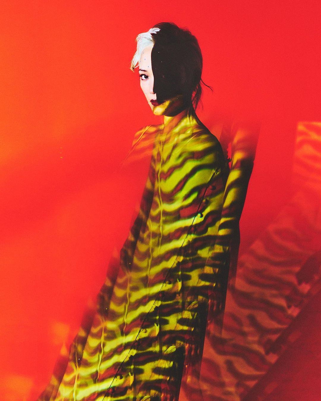 Woman poses in dress with tiger-stripe lighting in front of orange backdrop with half of her face in shadow in a studio setting.