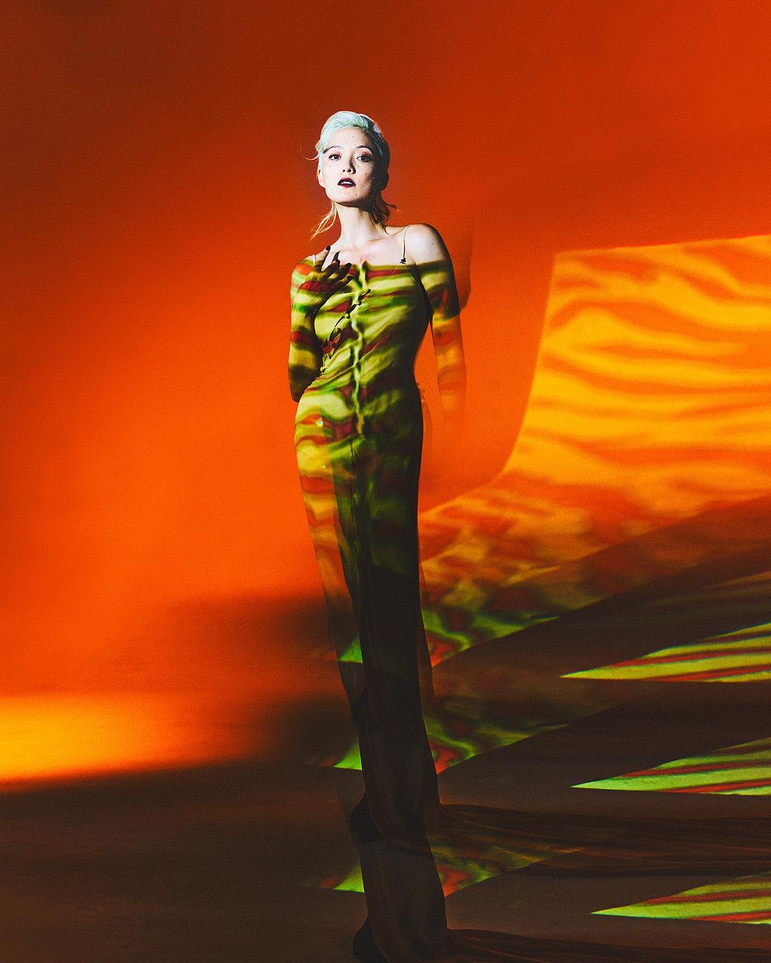 Woman in a striped off-shoulder dress stands in front of a surreal orange and yellow patterned backdrop in a studio setting.