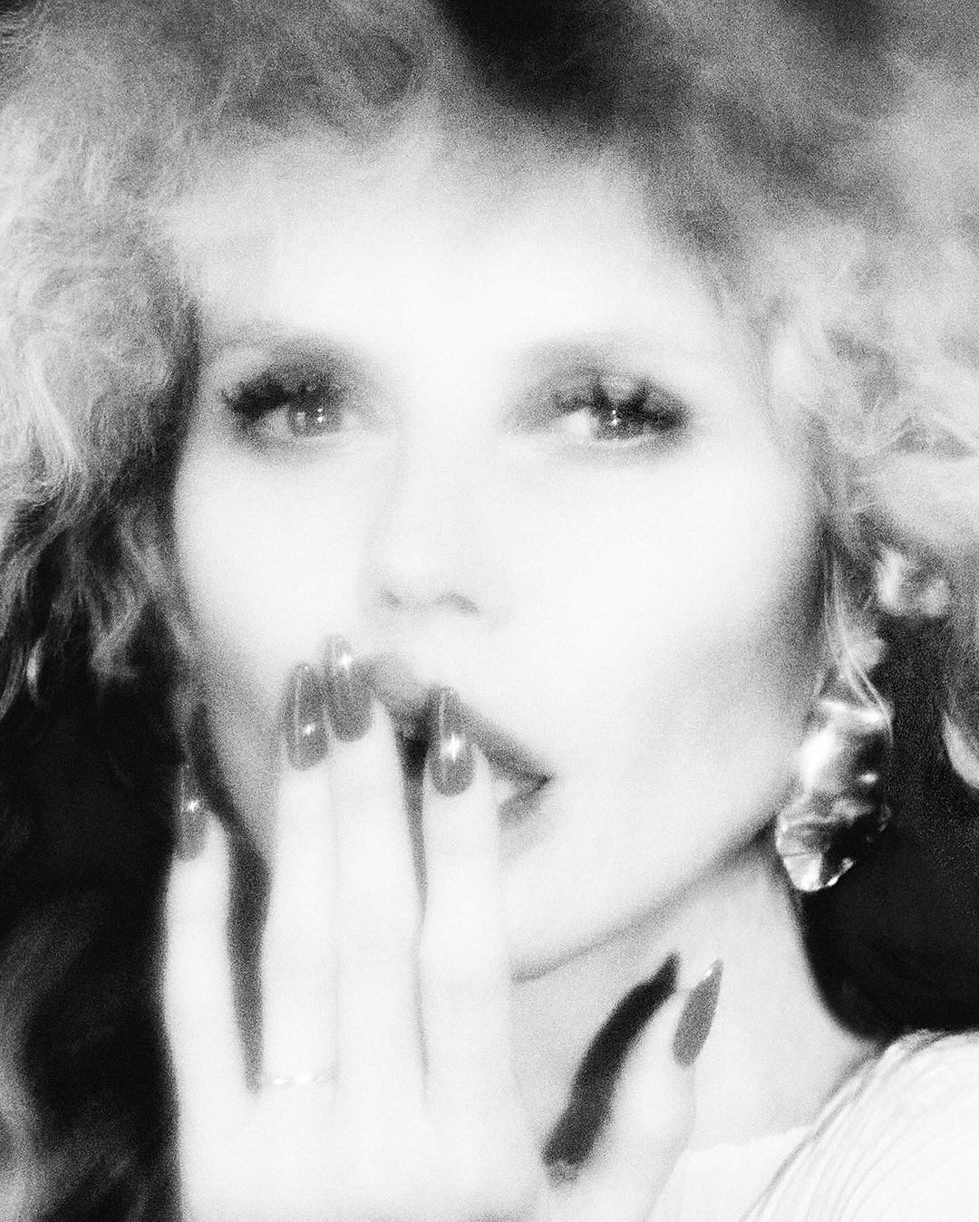 Close-up black and white portrait of a woman with manicured nails covering her mouth, wearing large earrings and a white top.