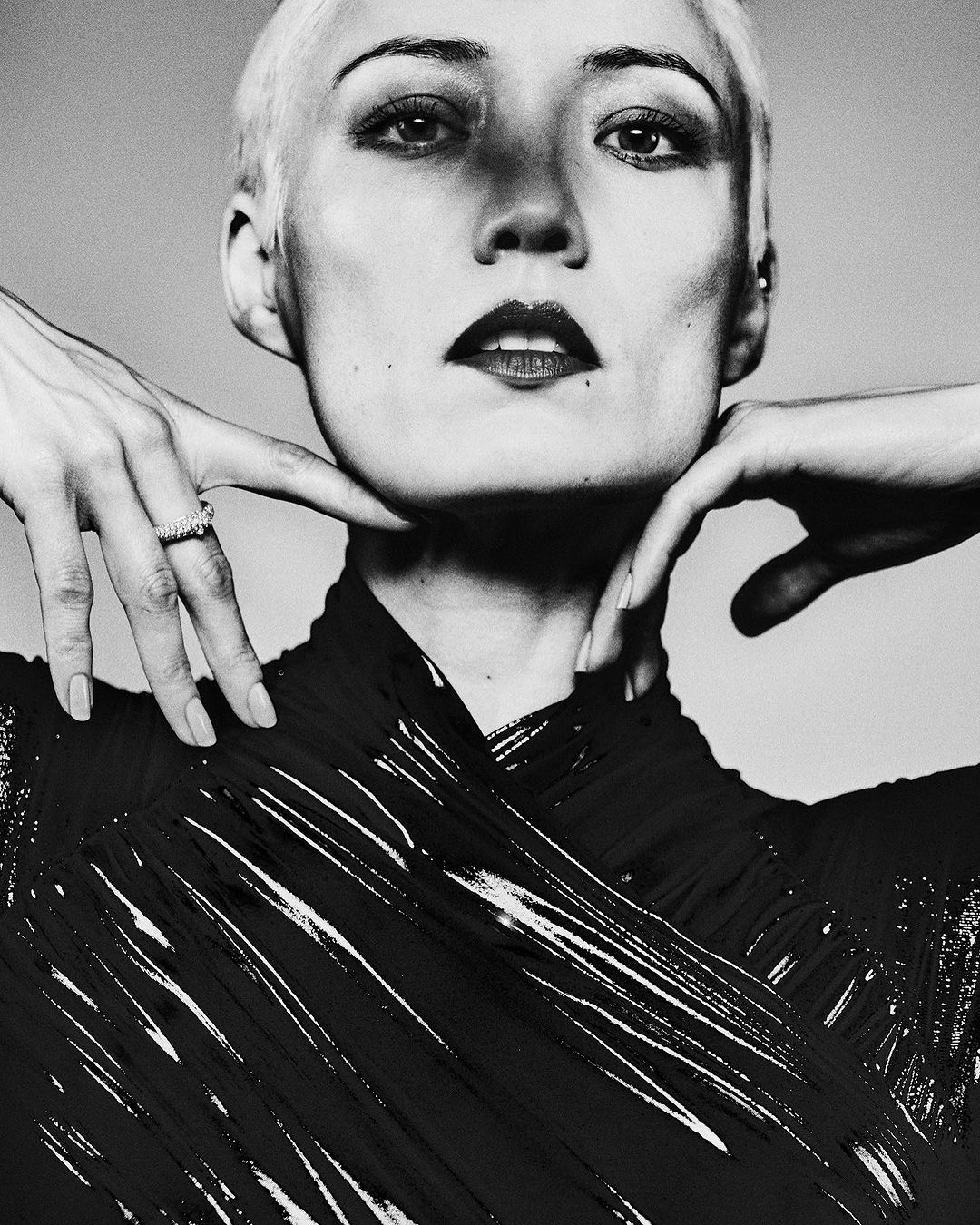Striking black and white portrait of a short-haired woman with hands framing her face, showcasing her makeup and unique style.