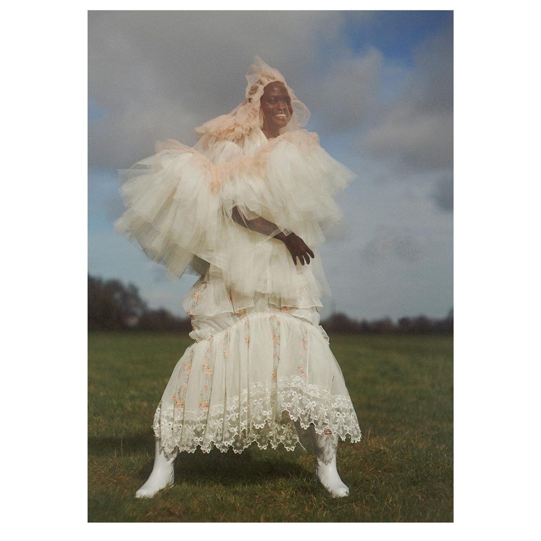 A lone figure stands in a field wearing a complex tiered couture dress and hood, under a cloudy sky, making a fashion statement.