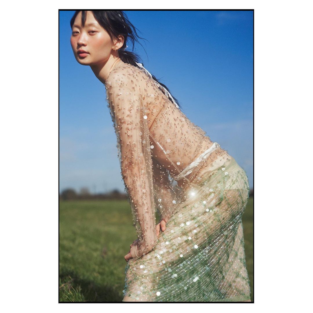 Asian woman in a sheer beaded dress poses in a sunlit meadow against a blue sky, showcasing a blend of fashion and nature.