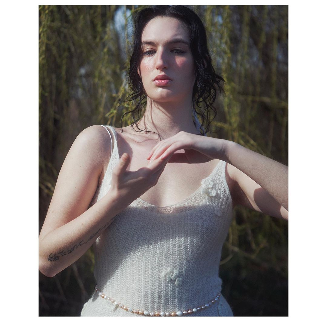 Portrait of a young woman with dark hair and fair skin wearing a light-colored knit dress outdoors with blurred green foliage background.