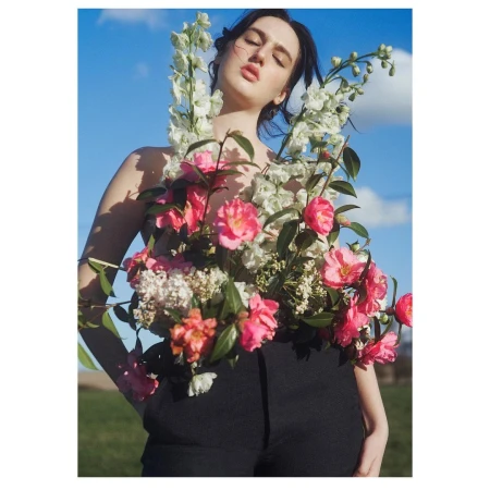 Woman poses outdoors wearing black pants and a large arrangement of pink and white flowers covering the upper part of her body.
