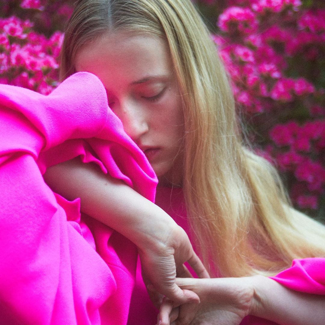 A blonde woman with long hair poses in front of a blurred nature backdrop, wearing a brown jacket and jeans in an eye-level medium shot.