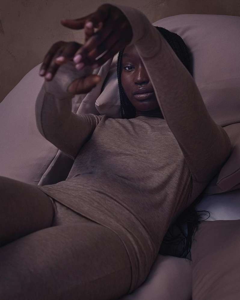 Portrait of Black woman in matching loungewear, lying on a bed with pillows, looking directly at the camera with hands framing her face.