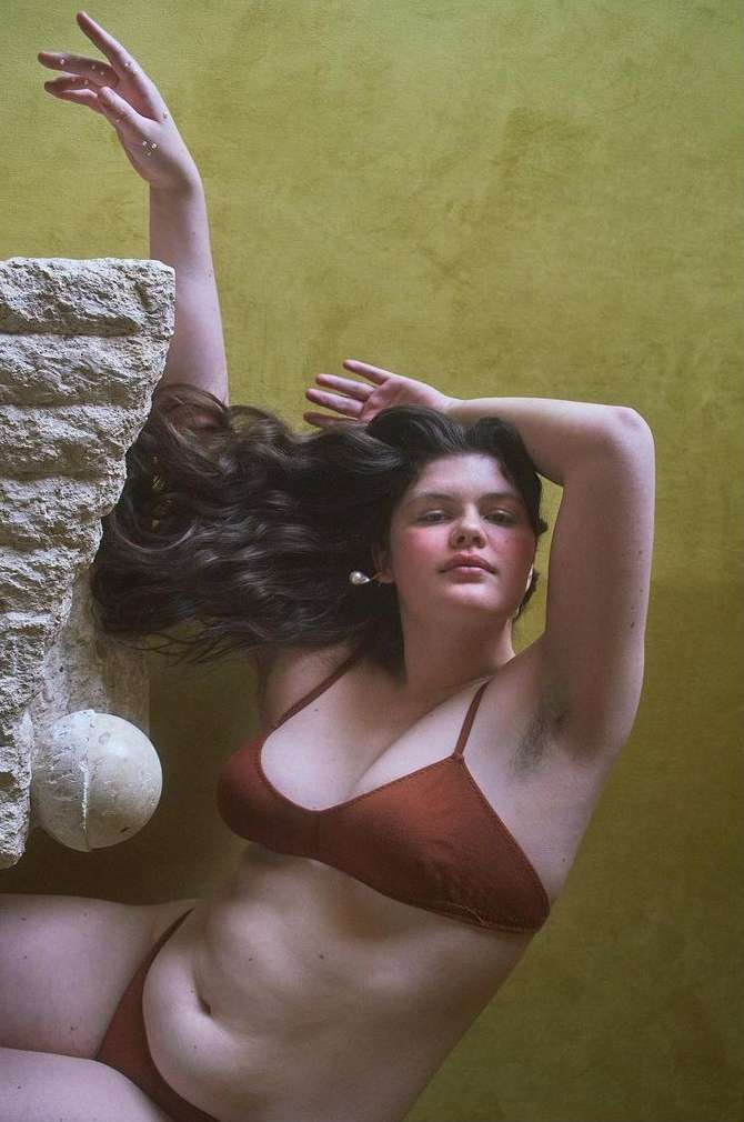 Woman in a brown bikini poses beside a textured architectural column with a white ball, against a mottled yellow-green background.