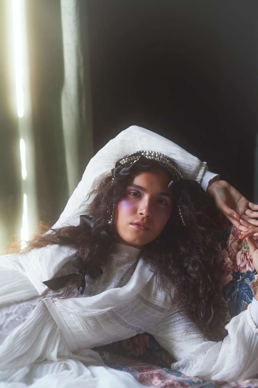 A woman with long curly hair wearing a white dress and jeweled headband poses on a floral-patterned surface in soft, diffused light.