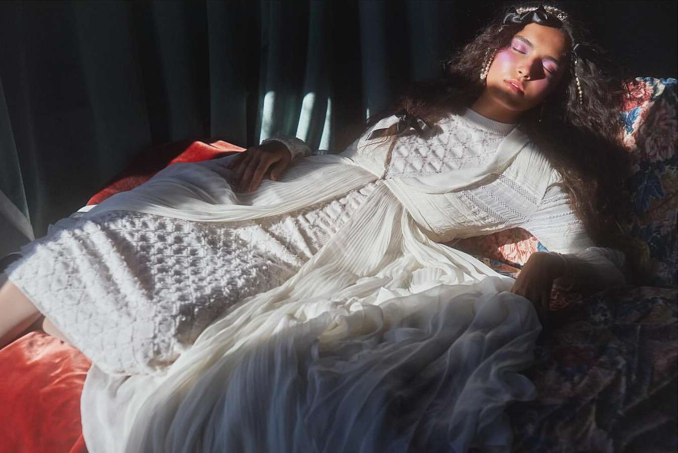 Young woman in white dress reclines on patterned fabric, illuminated by soft light, indoors. She wears pearl jewelry and has closed eyes.