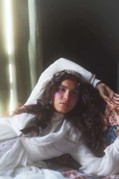 Woman in white vintage dress with dark curly hair posing on a floral couch in a sunlit room by Kristin Vicari