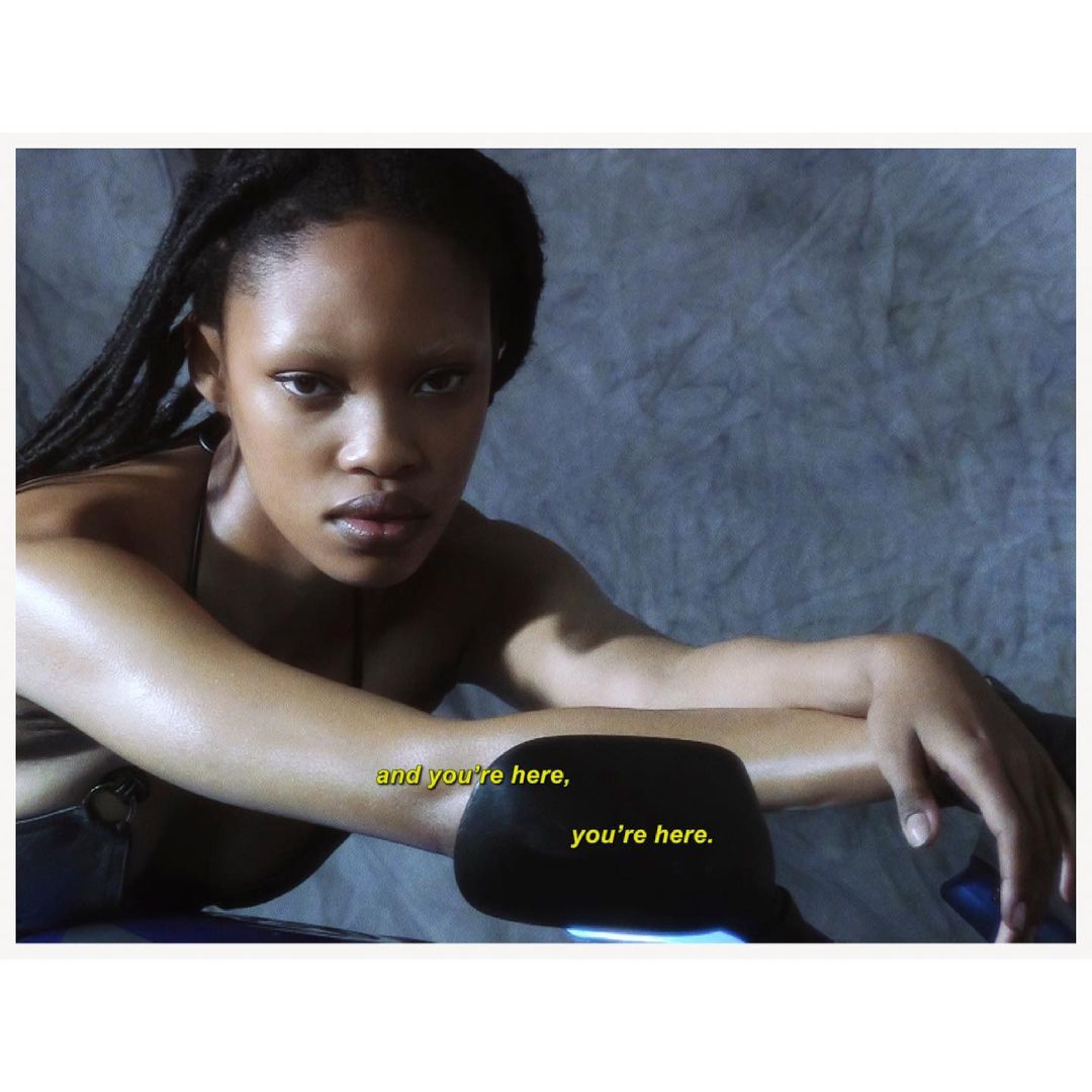 Portrait of a young Black woman with dreadlocks leaning on a motorcycle, soft studio lighting, grey textured backdrop, and visible text.