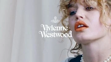Close-up studio shot of a model with blonde curly hair wearing Vivienne Westwood jewelry and bright orange lipstick.