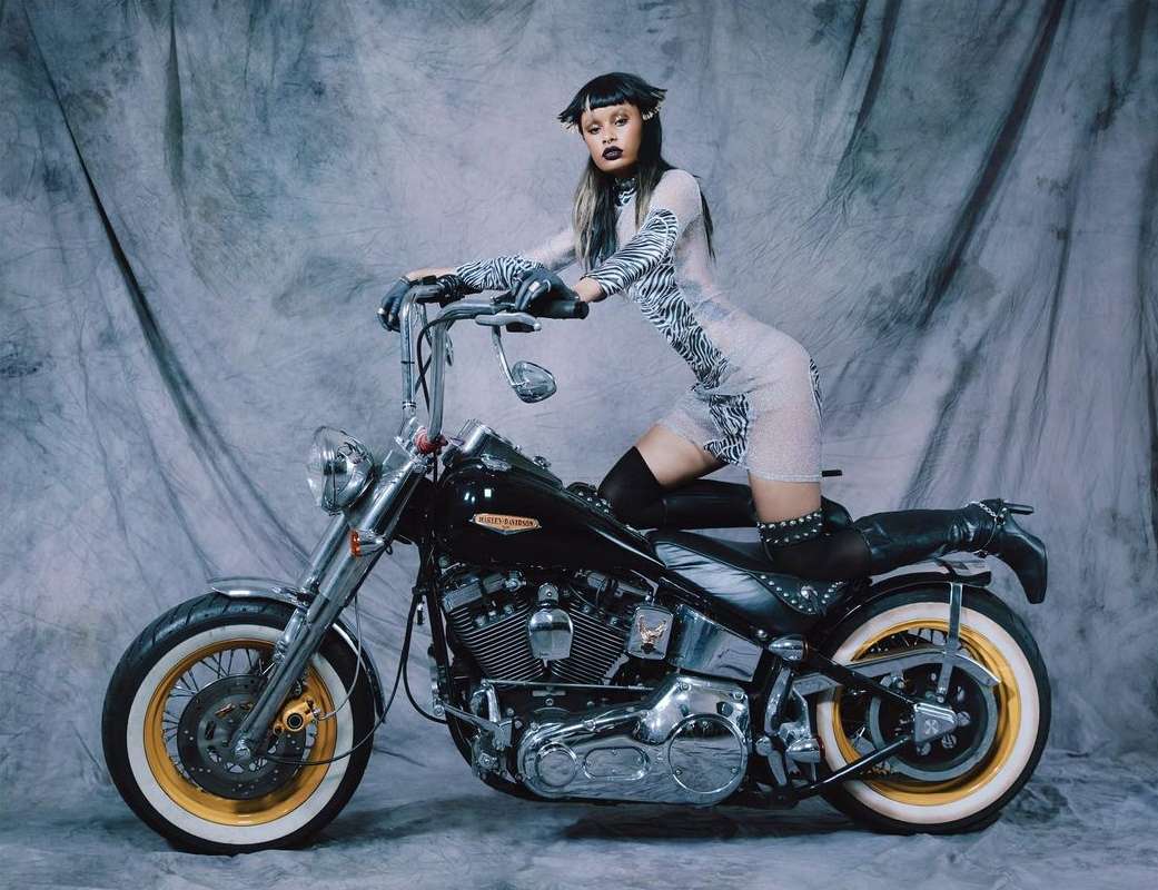 A woman poses on a black Harley Davidson motorcycle in a photo studio with a gray mottled backdrop, wearing a sheer zebra-print dress and black boots.