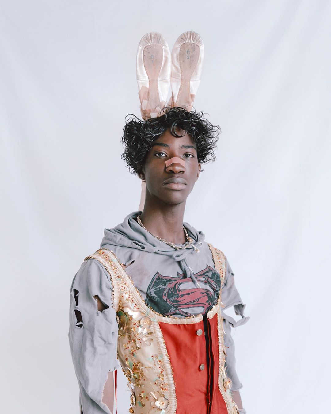 Portrait of a stylish Black adult wearing a hoodie and embellished vest with ballet pointe shoes balanced on their head in a studio setting.