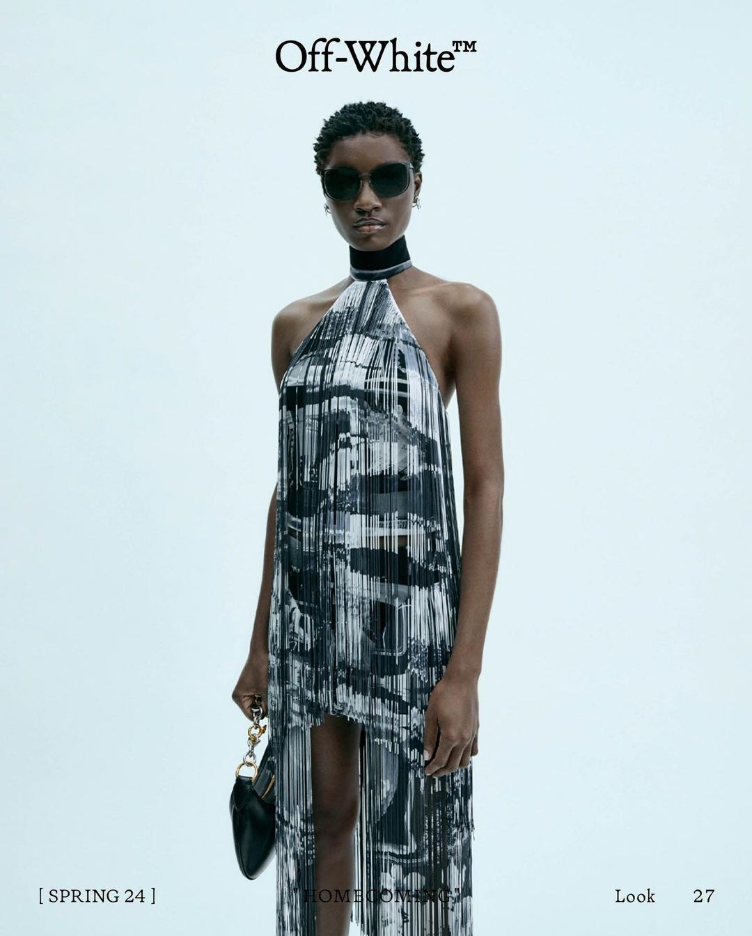 Model in printed halter dress and sunglasses holding a black handbag against a pale grey background