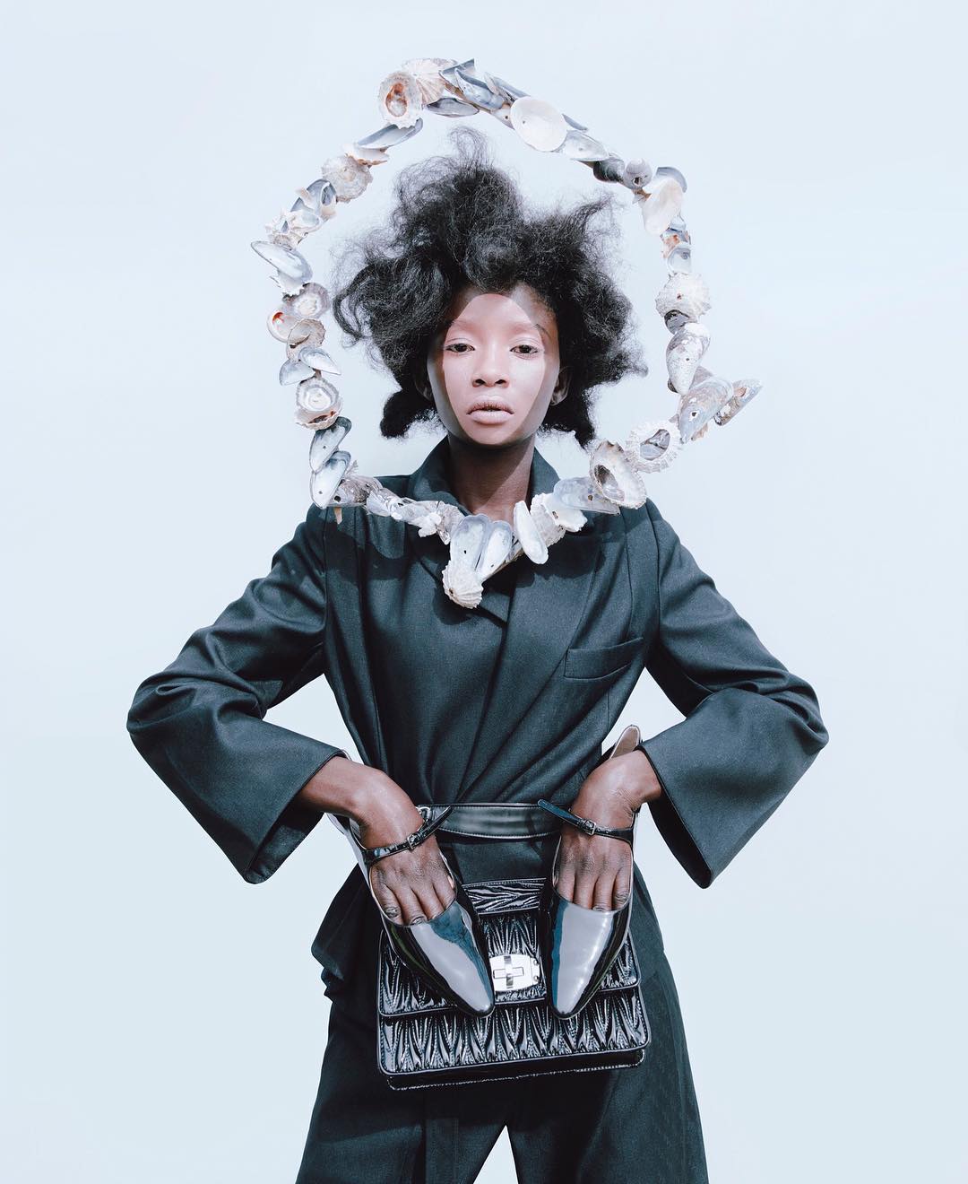 Fashion editorial shot of an adult female model wearing a dark suit, holding shoes and a purse in front of a white studio backdrop.
