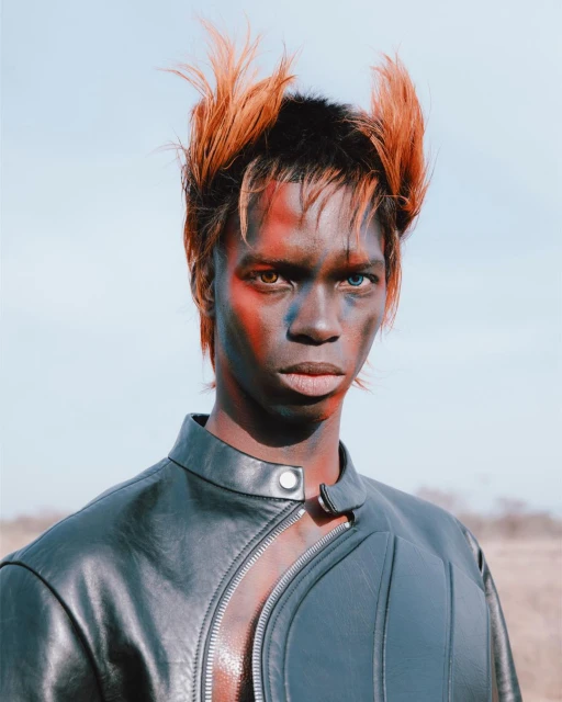 Close-up studio portrait of a person with dark skin, orange and black styled hair, and face paint, wearing a black leather jacket.