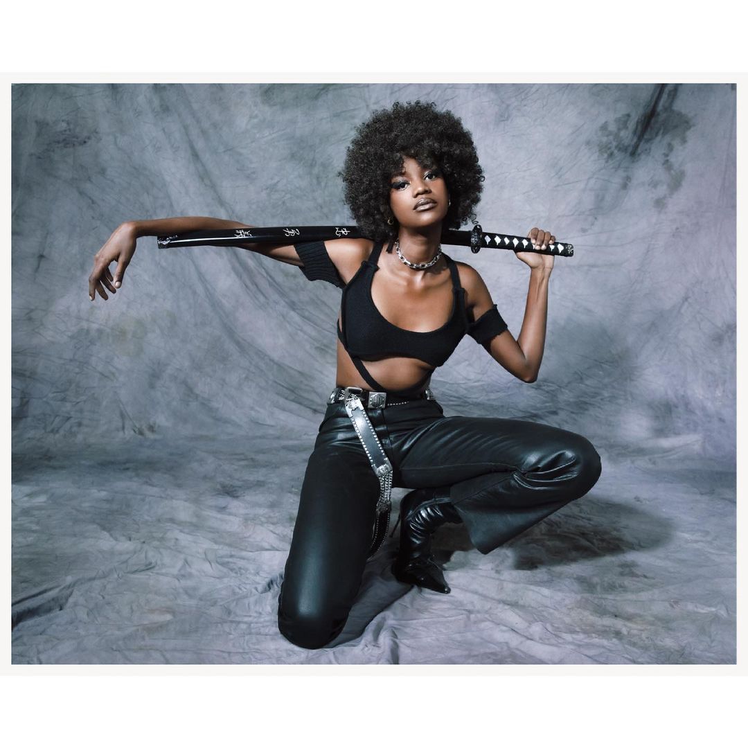 Attractive woman with afro hairstyle kneeling in stylish black outfit with a katana sword over shoulder, against gray textured backdrop.