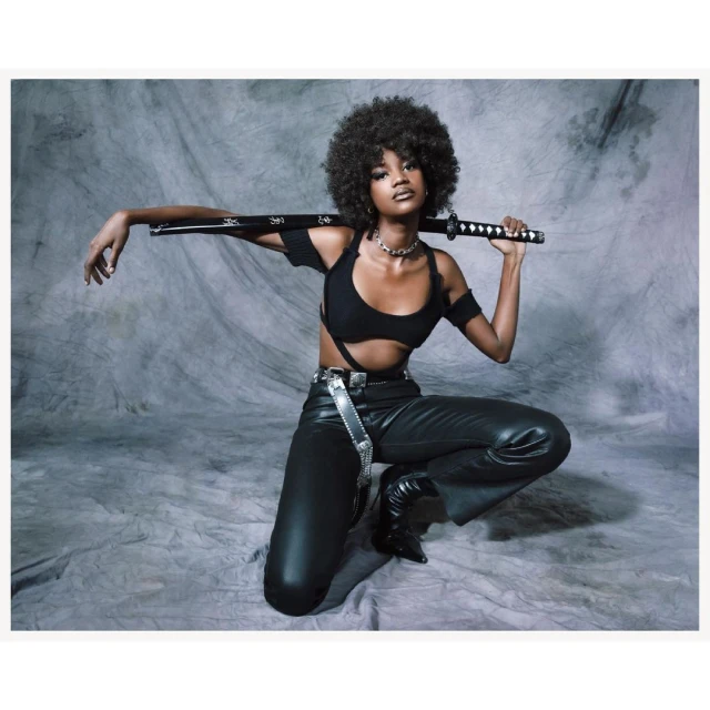 Attractive woman with afro hairstyle kneeling in stylish black outfit with a katana sword over shoulder, against gray textured backdrop.