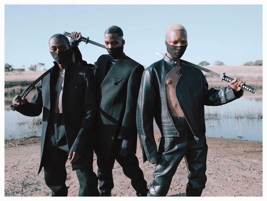 Three African male fashion models outdoors. They are wearing black ninja-style outfits and holding swords against a sunny landscape.
