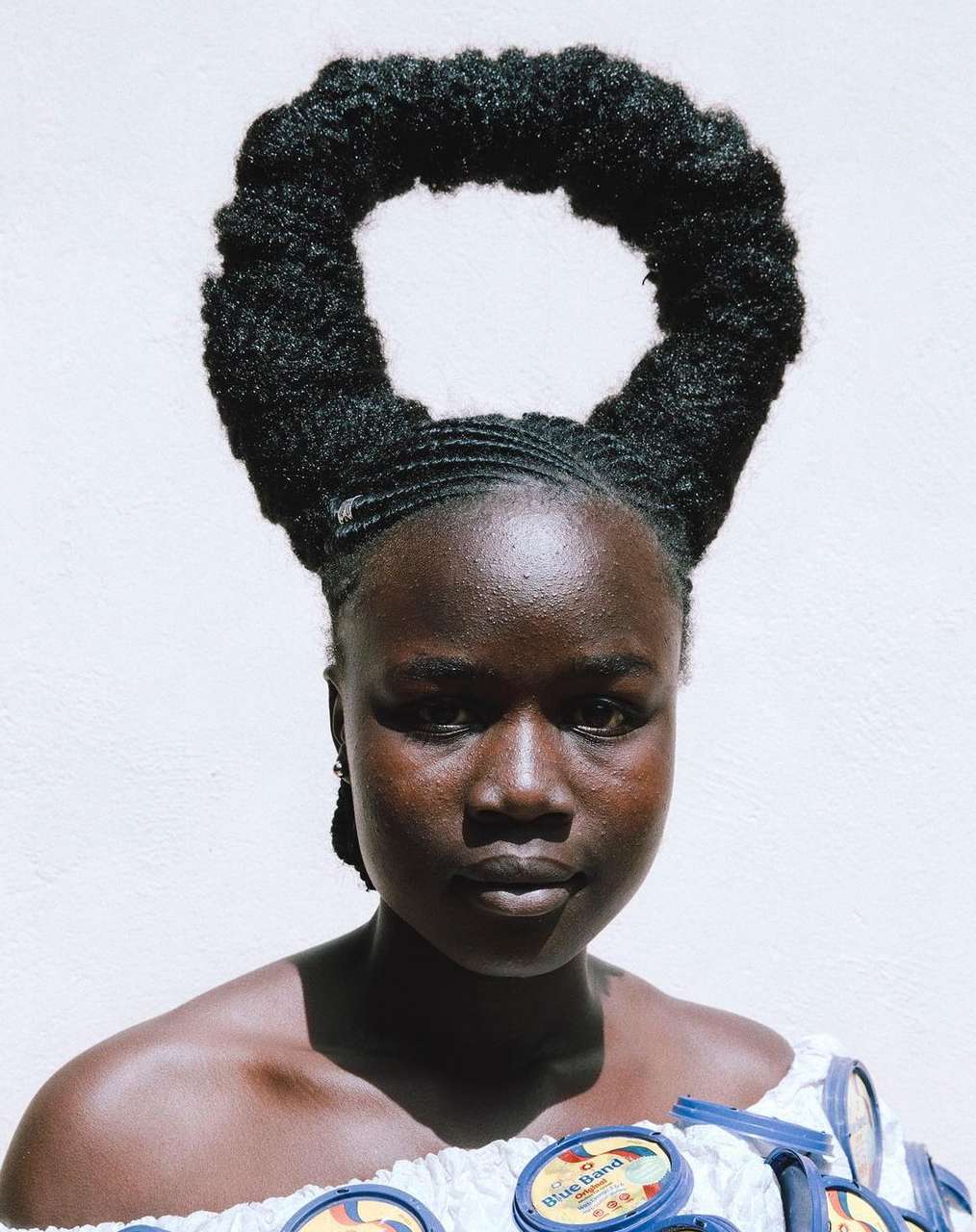 Portrait of a dark-skinned woman with unique afro hairstyle and patterned off-the-shoulder top against a plain white background.
