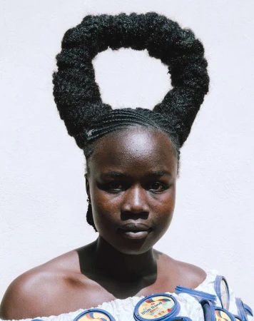 Portrait of a dark-skinned woman with unique afro hairstyle and patterned off-the-shoulder top against a plain white background.