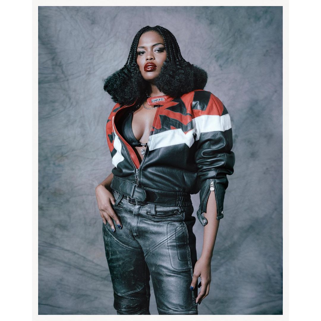 Fashion portrait of a woman in a leather jacket, bra top, and pants against a grey background, showcasing a bold and edgy style.