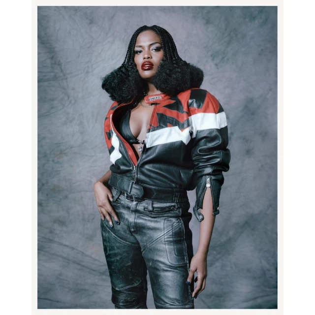 Fashion portrait of a woman in a leather jacket, bra top, and pants against a grey background, showcasing a bold and edgy style.