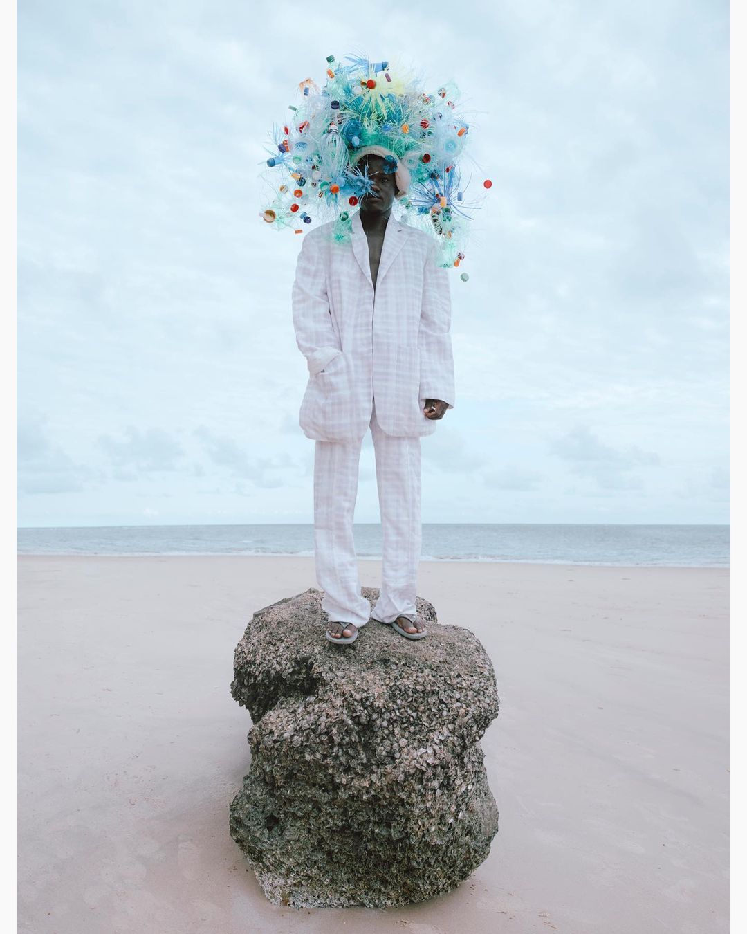 A person in a white suit standing on a rock at the beach wearing a sculptural headdress made of colorful plastic bottles.