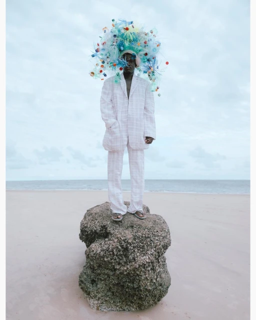 A person in a white suit standing on a rock at the beach wearing a sculptural headdress made of colorful plastic bottles.