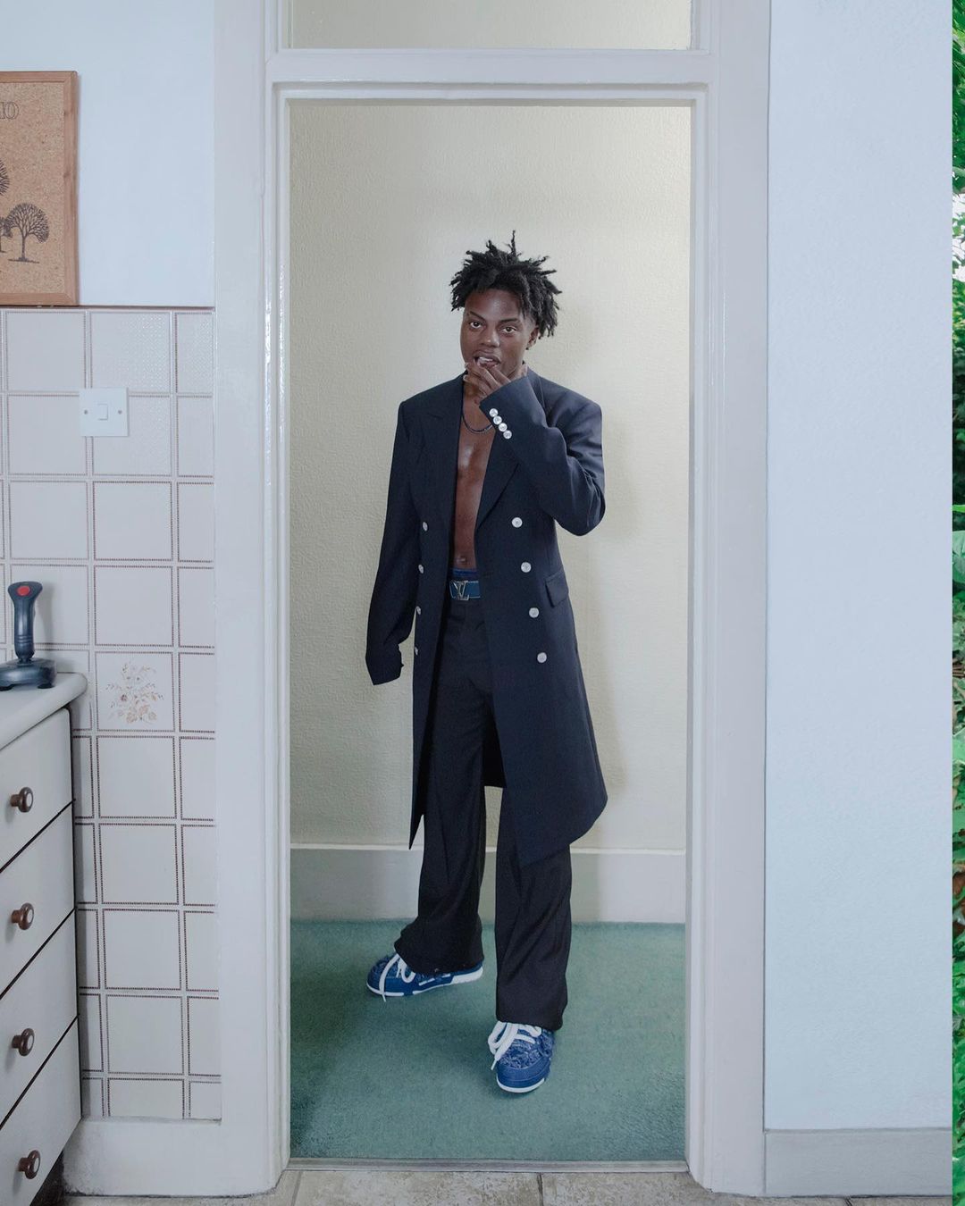 Black man standing in a doorway wearing a long dark coat over trousers and blue sneakers, photographed by Kristin-lee Moolman