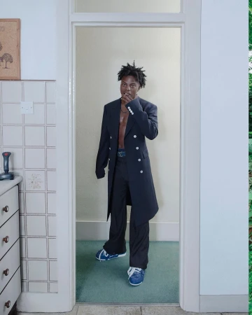 Black man standing in a doorway wearing a long dark coat over trousers and blue sneakers, photographed by Kristin-lee Moolman
