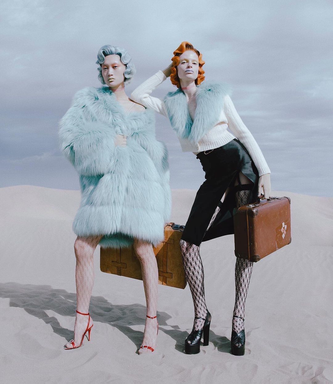 Two women in fashionable outfits stand in a desert-like landscape, carrying vintage suitcases, embodying a retro travel aesthetic.